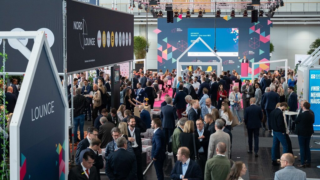 Innenansicht einer Messehalle aus mittlerer Distanz mit Besucherinnen und Besuchern im Gespräch sowie einzelnen Messeständen.