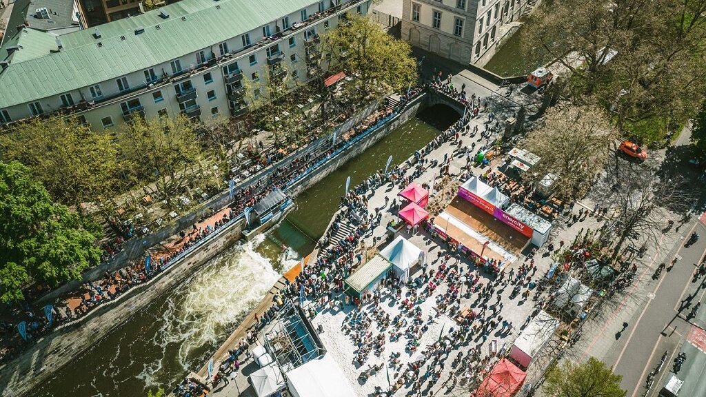 Ein Luftbild einer künstlich angelegten Surfwelle in der Innenstadt umgeben von Menschenmengen und Veranstaltungszelten.