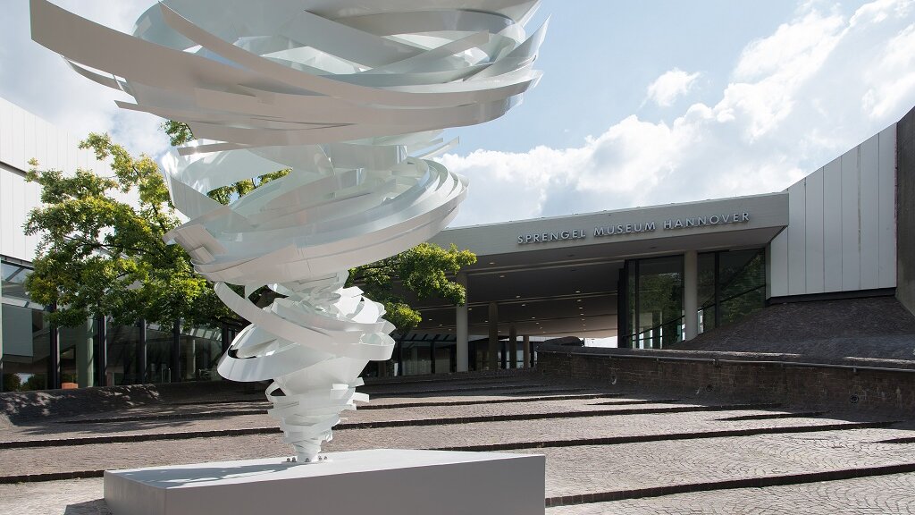 Vorderansicht des Sprengelmuseums Hannover von außen mit einer weißen Skulptur im Vordergrund.